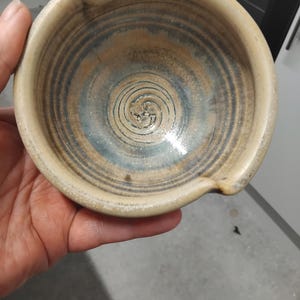May include: A round ceramic bowl with a beige exterior and a glazed interior featuring concentric blue and brown spiral patterns. The rim includes two small notches on opposite sides, designed for resting utensils or pouring liquids from the vessel.