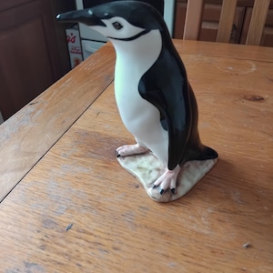 Könnte beinhalten: Eine Keramik-Pinguin-Figur mit schwarz-weißem Körper, stehend auf einem kleinen, unregelmäßigen Sockel. Der Pinguin hat einen schwarzen Schnabel und Füße. Die Figur steht auf einem Holztisch.