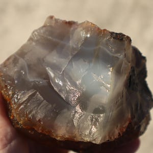 May include: A rough-cut, natural stone with a mix of translucent and opaque sections. The stone has a light blue and brown color palette, with a crystalline structure. The surface is uneven and textured, with areas that catch the light.