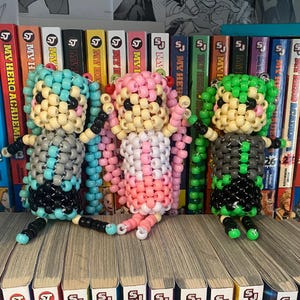 May include: Three beaded figures, each with a different color scheme. The figure on the left is blue, gray, and black. The middle figure is pink, white, and yellow. The figure on the right is green, gray, and black. The figures are standing on a shelf with a row of books behind them.