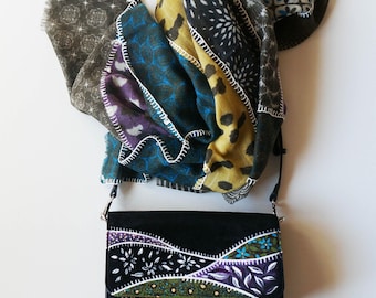 May include: A black crossbody bag with a flap featuring hand-painted floral and leaf patterns in white, purple, blue, and green. A multi-colored scarf with leopard print, geometric shapes, and floral motifs is draped behind the bag with white stitching.