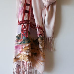 May include: A blush-colored scarf featuring a watercolor painting of the Golden Gate Bridge in San Francisco. The scarf has a fringed edge and is made of a soft, lightweight material. The bridge is depicted in shades of red and orange.