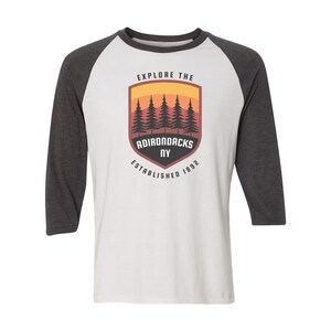 May include: A white and grey baseball tee shirt with a vintage-style logo featuring a sunset scene with pine trees and the text "Explore the Adirondacks NY Established 1892".