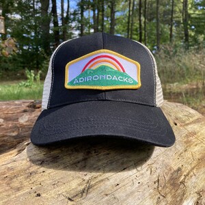 May include: Black trucker hat with a white mesh back and a curved brim. The front features a patch with the word "ADIRONDACKS" and a mountain scene with a rainbow. The hat is sitting on a log.