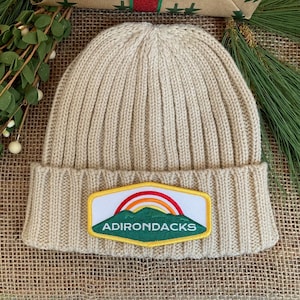 May include: A beige ribbed knit beanie hat with a rectangular patch featuring a rainbow over a mountain range and the word "ADIRONDACKS" in green. The hat is made of a soft, warm material, perfect for cold weather.