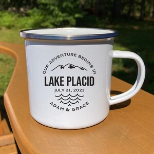 May include: White enamel camping mug with a black design that says "Our adventure begins in Lake Placid, July 21, 2021, Adam & Grace".