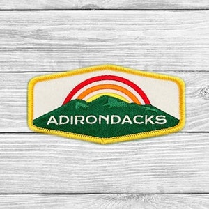 May include: Embroidered patch featuring the word "ADIRONDACKS" in white, set against a green mountain range and a rainbow. The patch has a yellow border and a cream-colored background. Ideal for jackets, bags, or other fabric items.