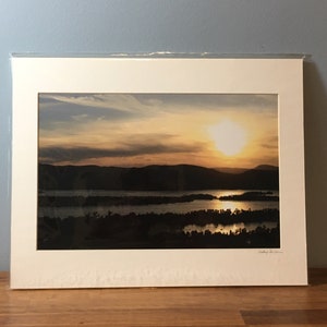 May include: A framed photograph of a sunset over a lake with islands. The sun is setting behind a mountain range in the distance. The sky is a mix of orange, pink, and blue.