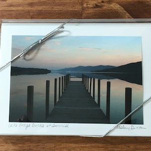 May include: A framed photograph of a wooden dock extending into a lake at sunrise. The dock is made of wooden planks and has a railing on either side. The lake is calm and the water is reflecting the sky. The mountains in the background are silhouetted against the sky. The caption on the photo reads "Lake George Docks at Sunrise" and is signed "Valley Dixon".