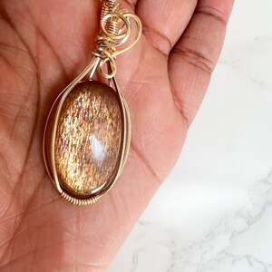 May include: A gold wire wrapped pendant featuring a large oval-shaped sunstone with a shimmering, iridescent finish.
