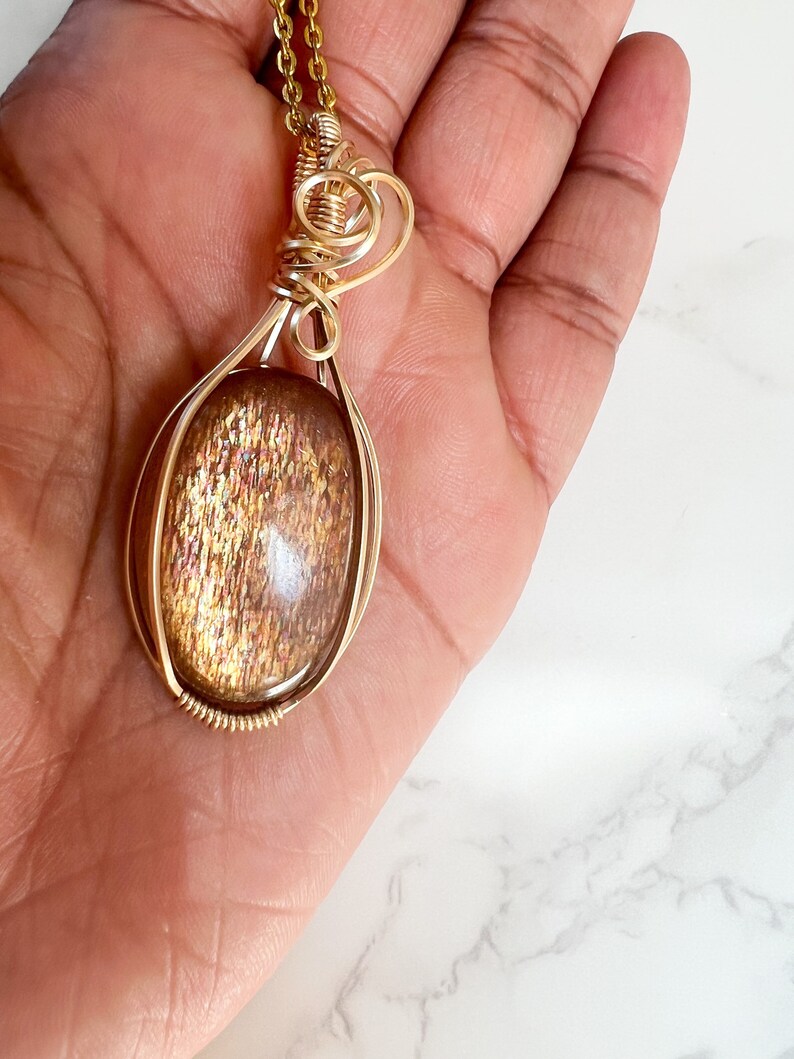 May include: A gold wire wrapped pendant featuring a large oval-shaped sunstone with a shimmering, iridescent finish.