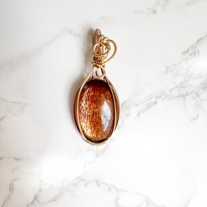 May include: A gold wire wrapped pendant featuring a sunstone cabochon. The pendant has a heart-shaped bail.