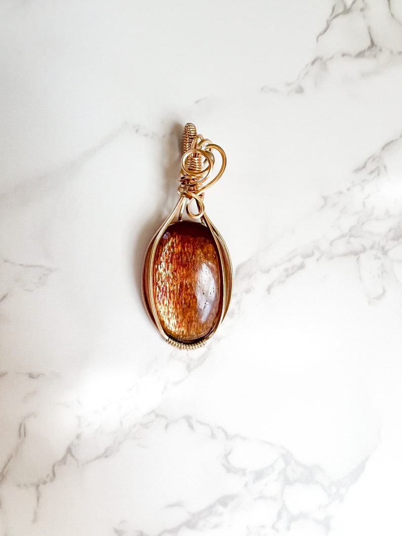 May include: A gold wire wrapped pendant featuring a sunstone cabochon. The pendant has a heart-shaped bail.