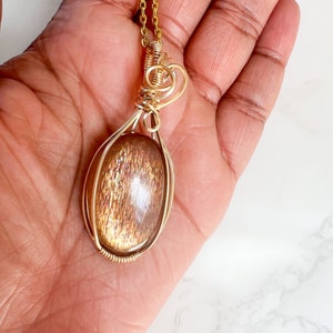 May include: A gold wire wrapped pendant featuring a sunstone gemstone. The pendant is oval shaped and has a gold chain.