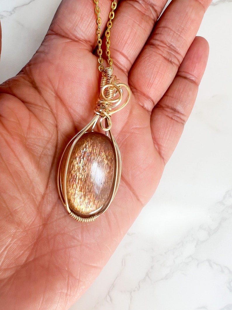 May include: A gold wire wrapped pendant featuring a sunstone gemstone. The pendant is oval shaped and has a gold chain.