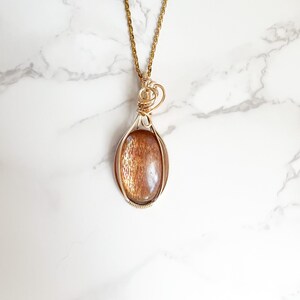 May include: Gold necklace with a sunstone pendant wrapped in gold wire. The pendant is oval shaped and has a reddish-brown color.