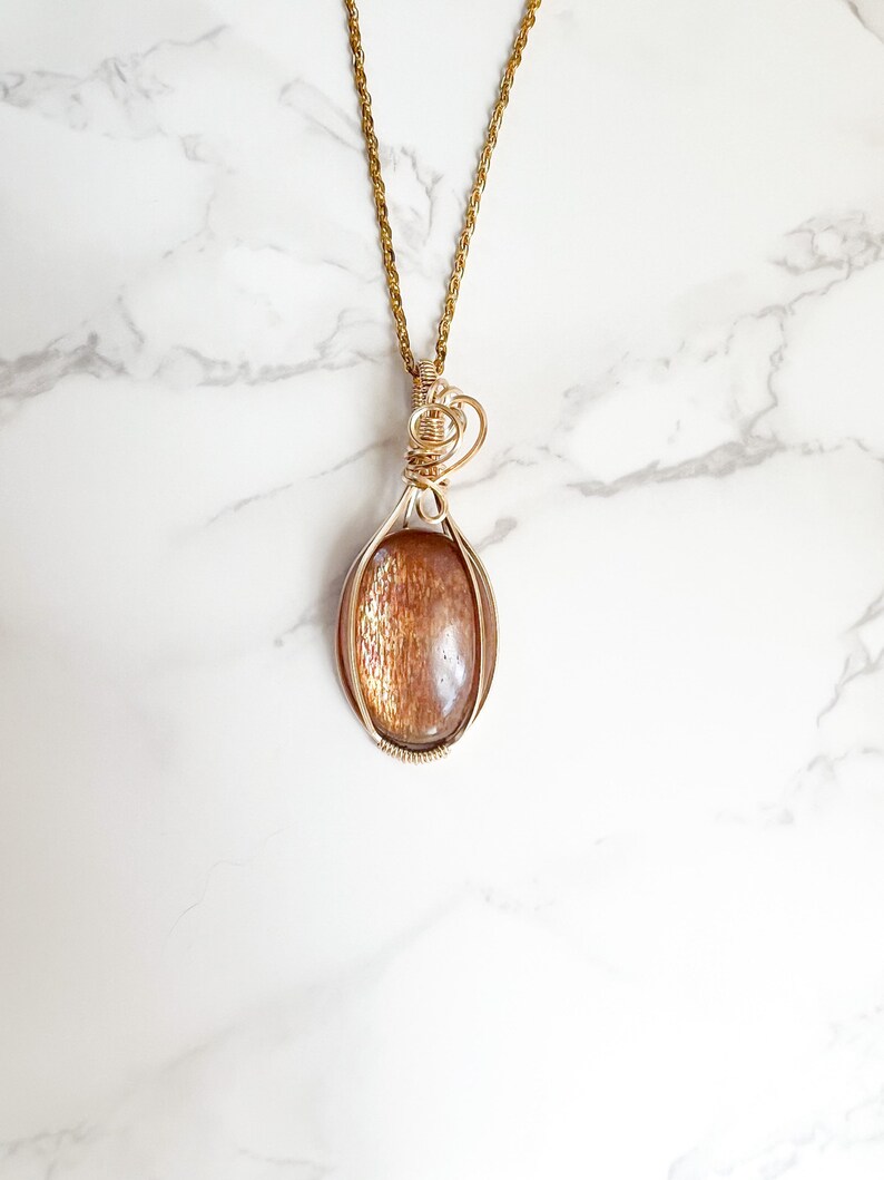 May include: Gold necklace with a sunstone pendant wrapped in gold wire. The pendant is oval shaped and has a reddish-brown color.
