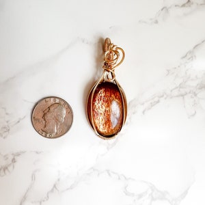 May include: A gold wire wrapped pendant featuring a brown sunstone gemstone. The pendant is oval shaped and is shown next to a US quarter for size comparison.