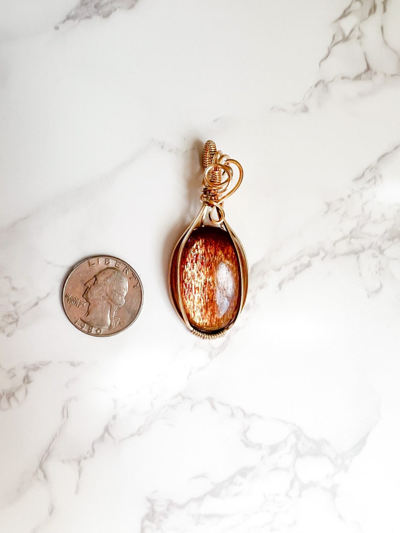May include: A gold wire wrapped pendant featuring a brown sunstone gemstone. The pendant is oval shaped and is shown next to a US quarter for size comparison.