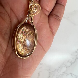 May include: Gold wire wrapped pendant featuring a large oval shaped sunstone with a shimmering, iridescent finish.