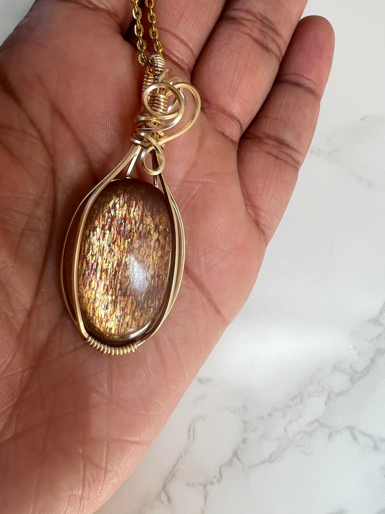 May include: Gold wire wrapped pendant featuring a large oval shaped sunstone with a shimmering, iridescent finish.