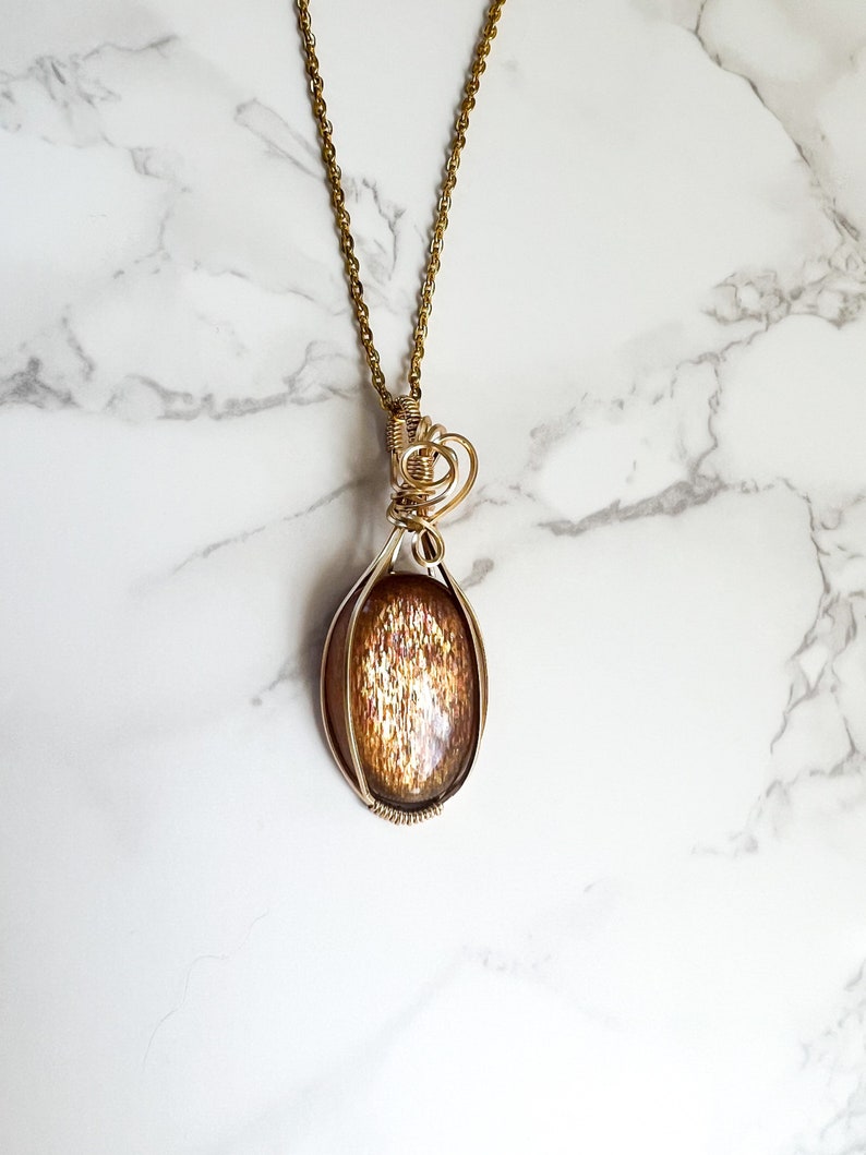 May include: A gold chain necklace with a pendant featuring a sunstone cabochon set in a wire wrapped oval frame.