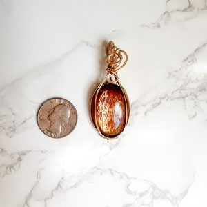 May include: Gold wire wrapped pendant featuring a brown sunstone gemstone. The pendant is approximately the size of a US quarter.