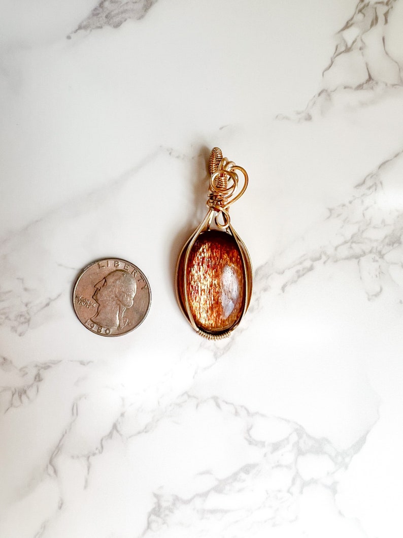 May include: Gold wire wrapped pendant featuring a brown sunstone gemstone. The pendant is approximately the size of a US quarter.