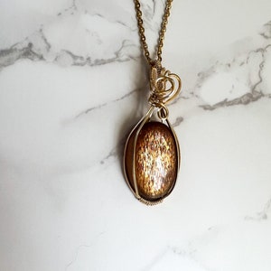 May include: Gold necklace with a sunstone pendant wrapped in wire. The sunstone is oval shaped and has a shimmery, iridescent finish.