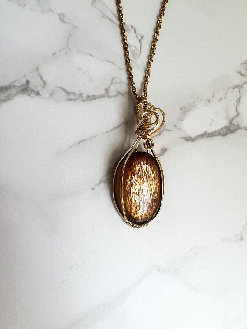 May include: Gold necklace with a sunstone pendant wrapped in wire. The sunstone is oval shaped and has a shimmery, iridescent finish.