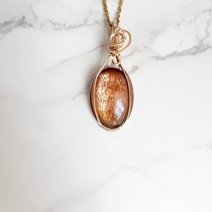 May include: Gold wire wrapped pendant featuring a sunstone gemstone. The pendant is oval shaped and has a wire bail for attaching to a chain.