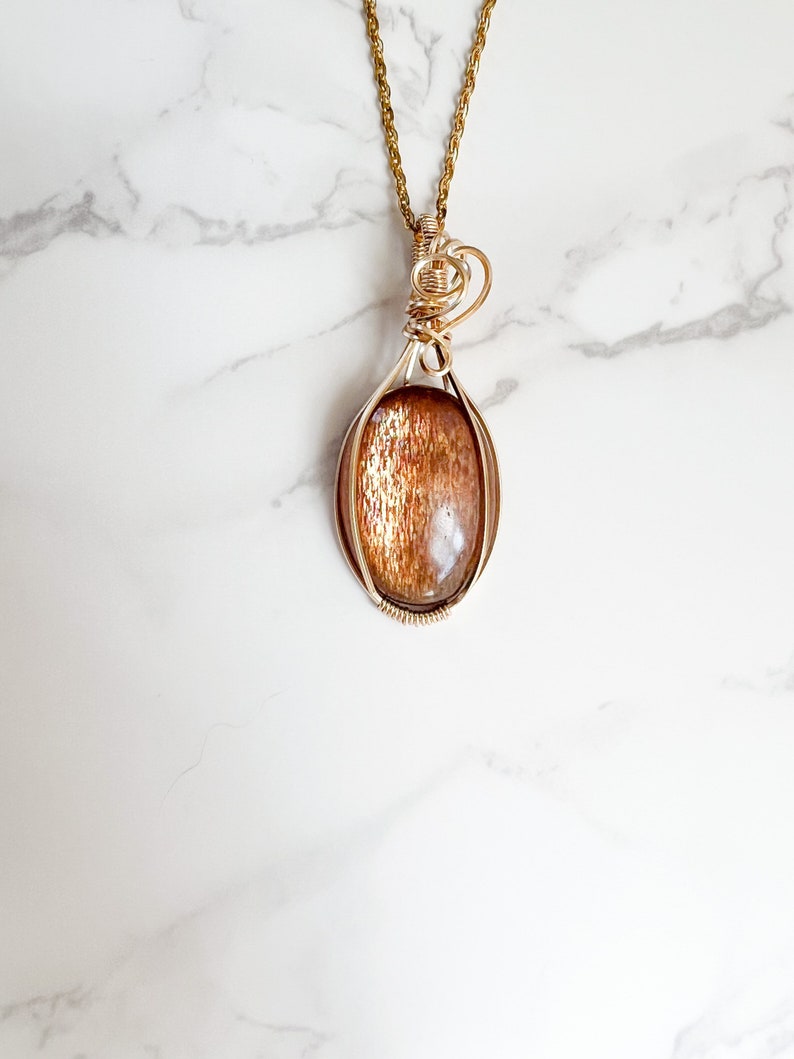 May include: Gold wire wrapped pendant featuring a sunstone gemstone. The pendant is oval shaped and has a wire bail for attaching to a chain.