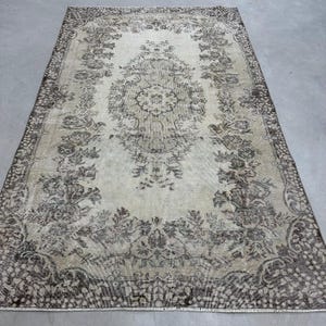 Puede incluir: Una alfombra rectangular de estilo vintage con un intrincado diseño floral. La alfombra presenta un diseño de medallón central y un borde con motivos florales. La paleta de colores incluye tonos beige, marrón y gris, creando una apariencia envejecida y antigua.