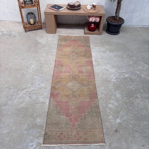 May include: A long, rectangular runner rug featuring a faded geometric pattern in shades of dusty pink, muted gold, and olive green. The design includes a central diamond motif with intricate borders and short, light-colored fringe at both ends.
