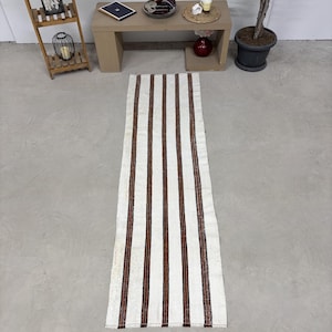 May include: A long, rectangular runner rug featuring a repeating pattern of vertical dark brown stripes set against a cream-colored background. The textile has a woven texture and finished edges, extending across the floor in a straight orientation.