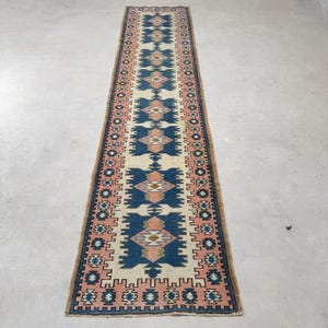 May include: A long, narrow runner rug featuring a repeating geometric pattern in shades of deep blue, soft pink, and cream. The design includes central diamond motifs and intricate border detailing, creating a symmetrical layout across the entire length.