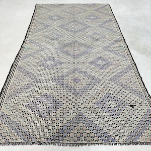 Puede incluir: Una alfombra rectangular con un patrón geométrico en tonos azules, crema y negro. La alfombra tiene un borde negro con flecos en los extremos cortos. El patrón consiste en líneas verticales y pequeñas formas de diamante.