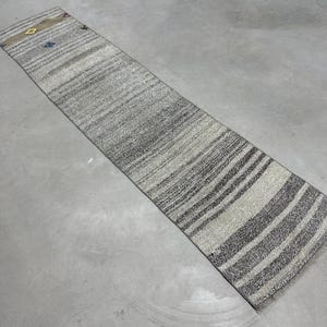 May include: A long, narrow, handwoven rug with a striped pattern in shades of gray and beige. The rug has a rectangular shape with a fringed edge. Small decorative elements in blue, yellow, and red are visible near the top.