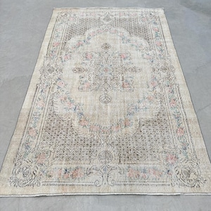 Peut inclure: Un tapis rectangulaire orné d'un médaillon floral central dans des tons rose et bleu sur un fond beige. Le tapis présente une bordure en treillis marron foncé avec des motifs floraux complexes et des volutes sur toute la surface.