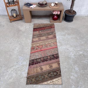 May include: A long, rectangular runner rug featuring a patchwork design of horizontal bands in muted tones of red, brown, beige, and gray. The rug displays various intricate geometric and floral patterns across its surface, creating a textured appearance.