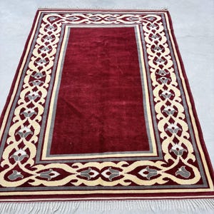May include: A rectangular burgundy rug with a cream and gray floral border. The rug has a central burgundy field and a cream-colored border with gray floral accents. The rug has a fringe on the short ends.