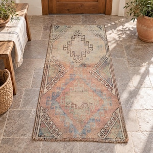 May include: A rectangular runner rug featuring a faded, distressed pattern in shades of peach, blue, and cream. The design includes central diamond motifs and geometric borders with a short fringe along the top and bottom edges of the textile.