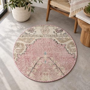 May include: A round area rug featuring a central pink medallion surrounded by cream and beige floral patterns with black outlines. The rug has a distressed texture and a pink stitched border, displaying a symmetrical design on a flat surface.