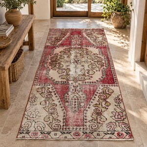 May include: A rectangular runner rug featuring a central medallion design in shades of red, cream, and muted green. The rug displays intricate geometric and floral patterns with a distressed, weathered texture throughout the woven fabric surface.