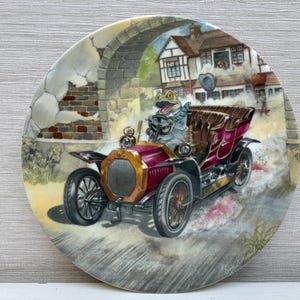 Könnte beinhalten: Dekorative Platte mit einer skurrilen Illustration eines Frosches, der ein Oldtimer-Bordeaux-Auto fährt. Der Frosch trägt einen Schal und fährt durch eine Stadt mit einem steinernen Torbogen. Die Platte hat einen goldenen Rand.