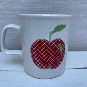 Könnte beinhalten: Weiße Keramiktasse mit einem roten Apfel-Design. Der Apfel hat einen grünen Stiel und ein Blatt und ein rot-weißes Karomuster.