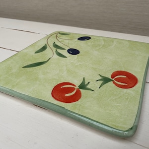 May include: Square green ceramic trivet with a painted design of olive branches, black olives, and red tomatoes.