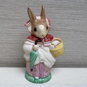 May include: A ceramic rabbit figurine wearing a white dress, red shawl, and bonnet. It holds a green umbrella and a yellow basket. The base is green with purple and pink details. The figurine is approximately 18 cm tall.