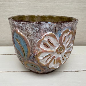 Könnte beinhalten: Ein weißer Keramiktopf mit einem braunen, blauen und goldenen Blumendesign. Der Topf hat einen leicht unebenen Rand und eine strukturierte Oberfläche.