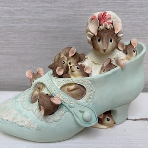 Puede incluir: Una figura de cerámica en forma de zapato azul claro con un grupo de ratones marrones. Un ratón más grande lleva un gorro blanco con un detalle rosa. Varios ratones pequeños están metidos en el zapato, incluido uno bajo el tacón.